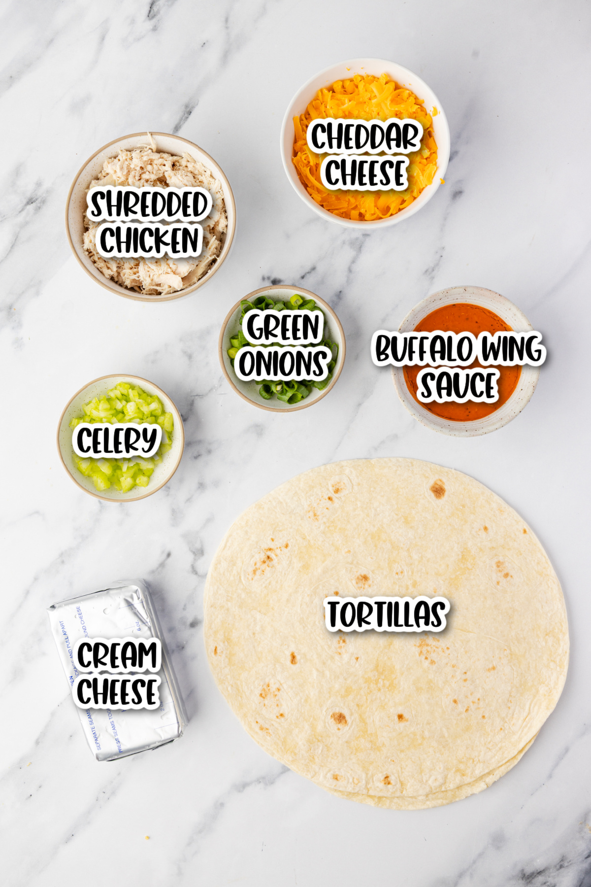 Ingredients for buffalo chicken pinwheels arranged on a white surface, including tortillas, cream cheese, celery, shredded chicken, green onions, cheddar cheese, and buffalo wing sauce.