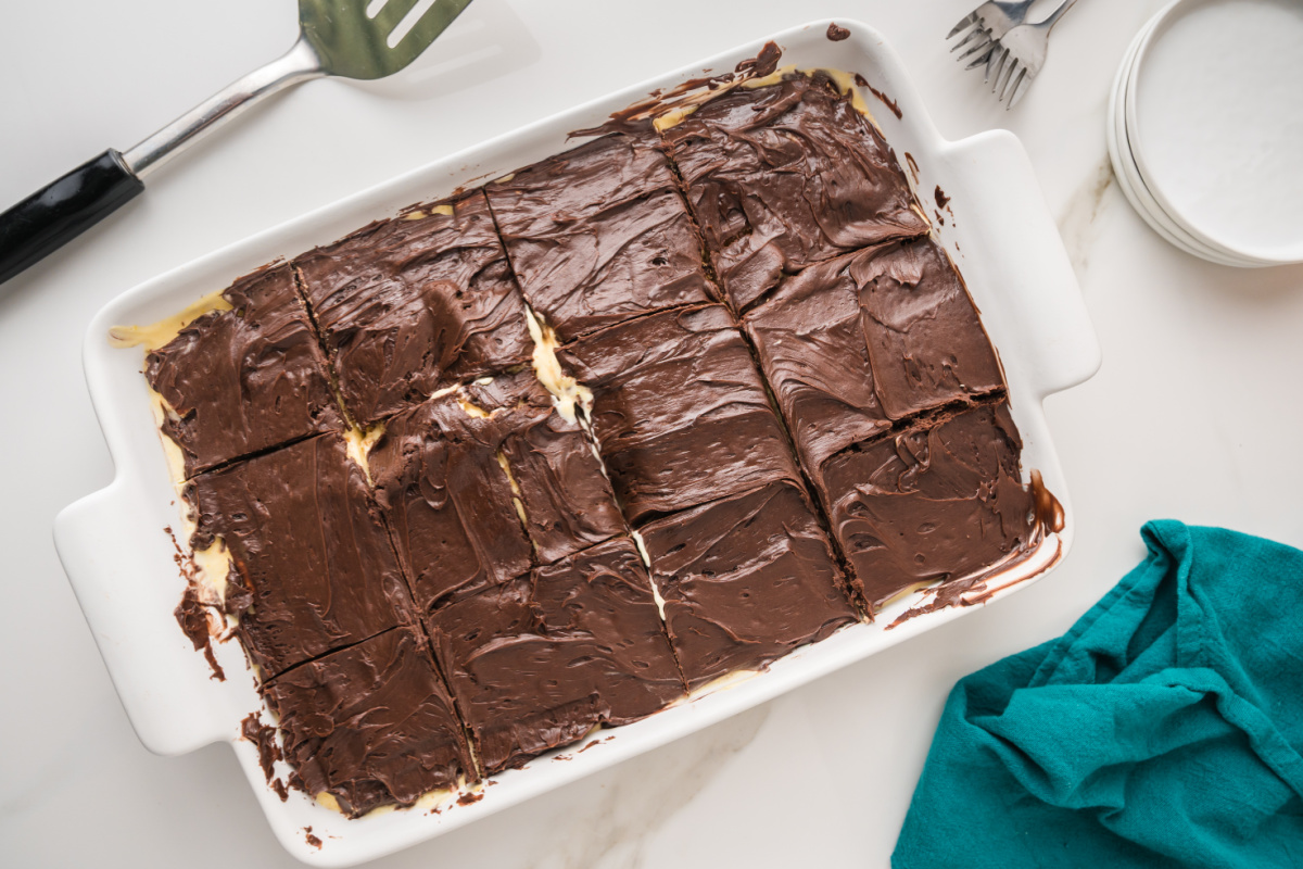 A baking dish of chocolate-frosted dessert bars cut into squares, with a spatula, forks, and a teal cloth nearby on a white surface.