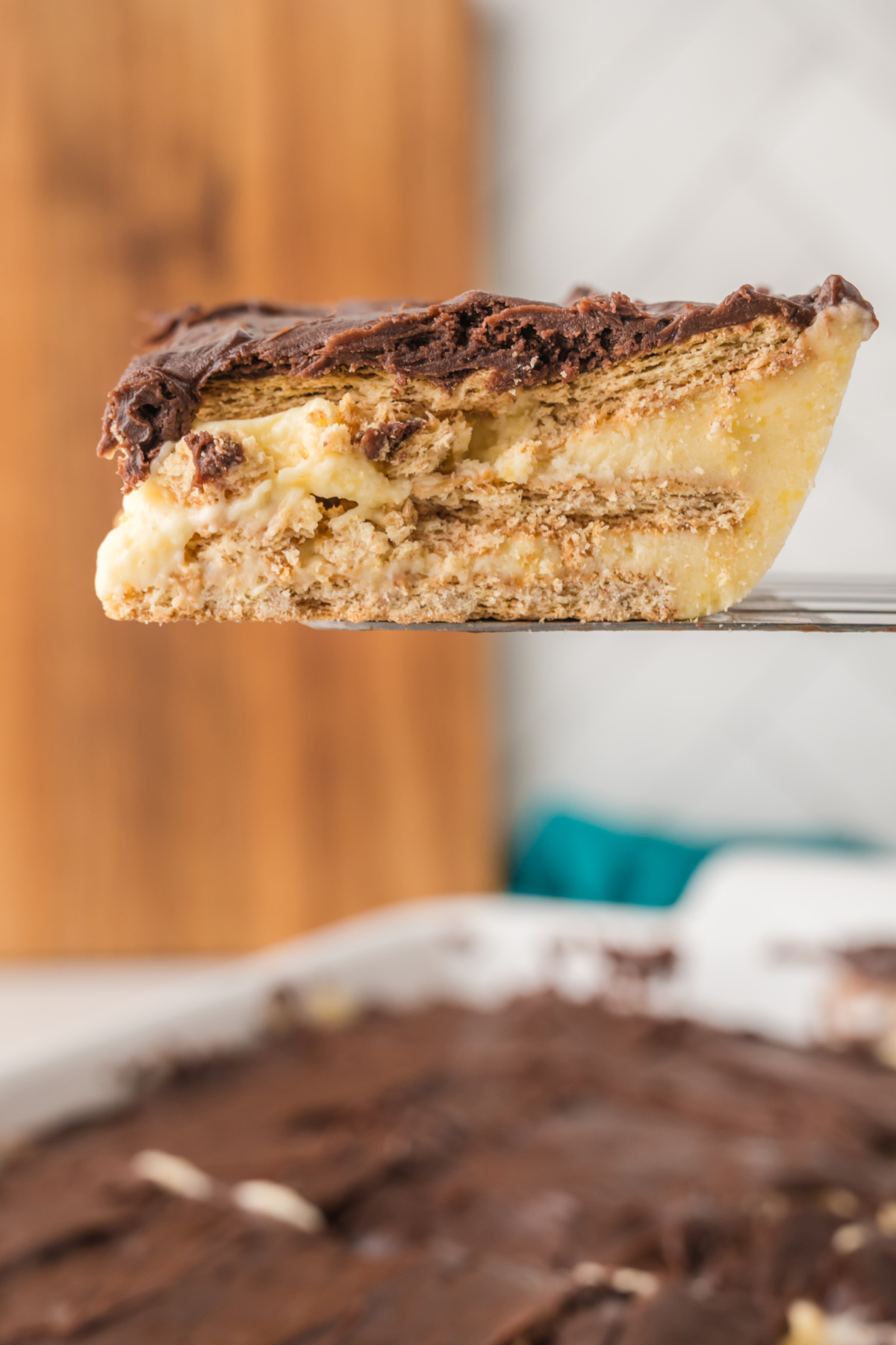 A close-up of a slice of layered dessert with a chocolate topping and a creamy, biscuit-filled base held above a larger pan of the same dessert.