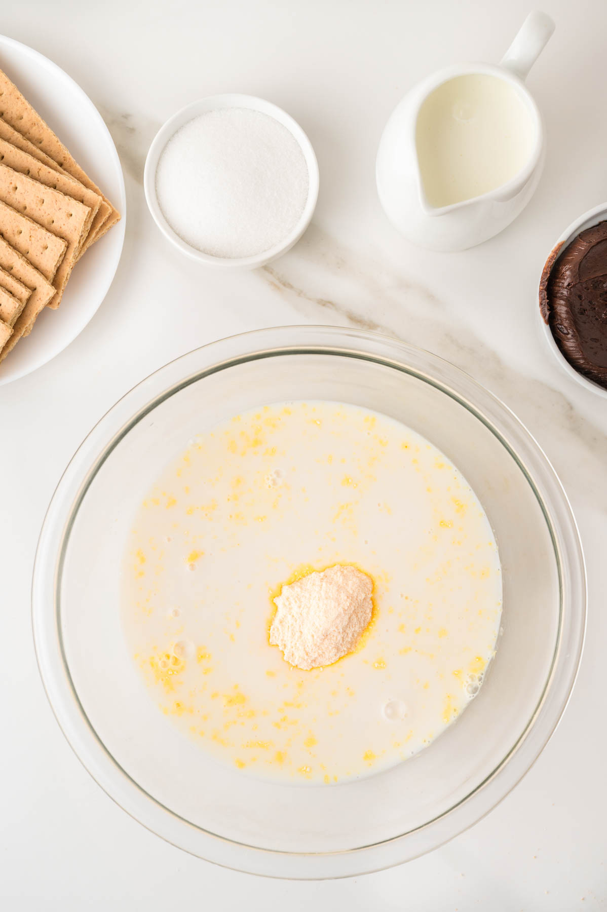 A glass bowl with pudding and milk. Surrounding it are sugar, graham crackers, a pitcher of milk, and a bowl of chocolate.