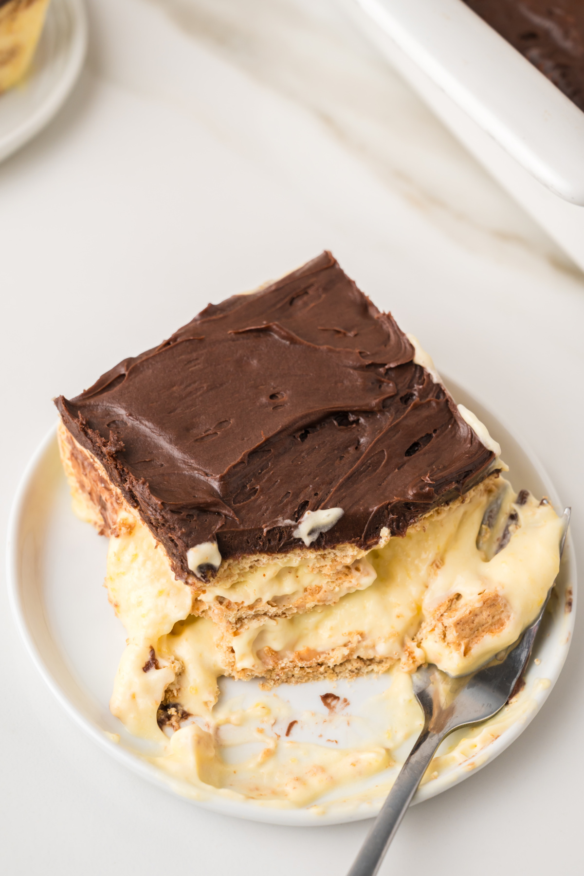 A slice of layered dessert with chocolate topping, creamy filling, and graham crackers, served on a white plate with a fork.