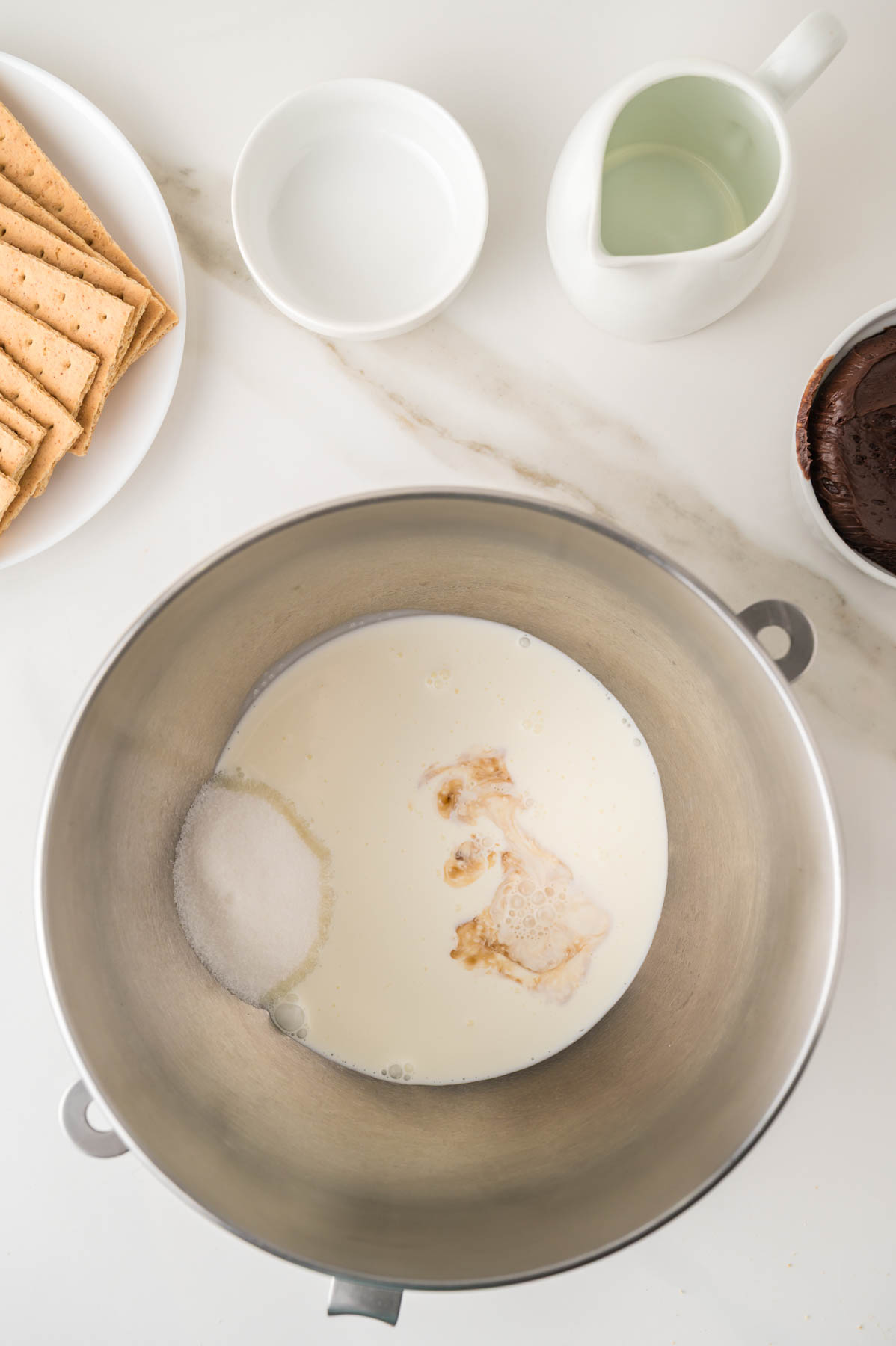 A metal mixing bowl with cream, sugar, and vanilla; nearby are graham crackers on a plate, a small bowl, a pitcher, and a bowl of chocolate.