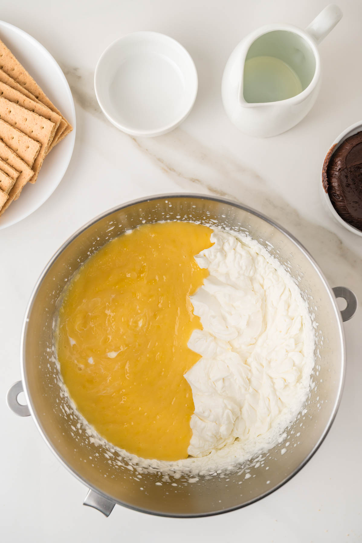 A mixing bowl with whipped cream and yellow pudding mix side by side, surrounded by graham crackers, a small pitcher, a bowl of water, and chocolate filling on a white countertop.