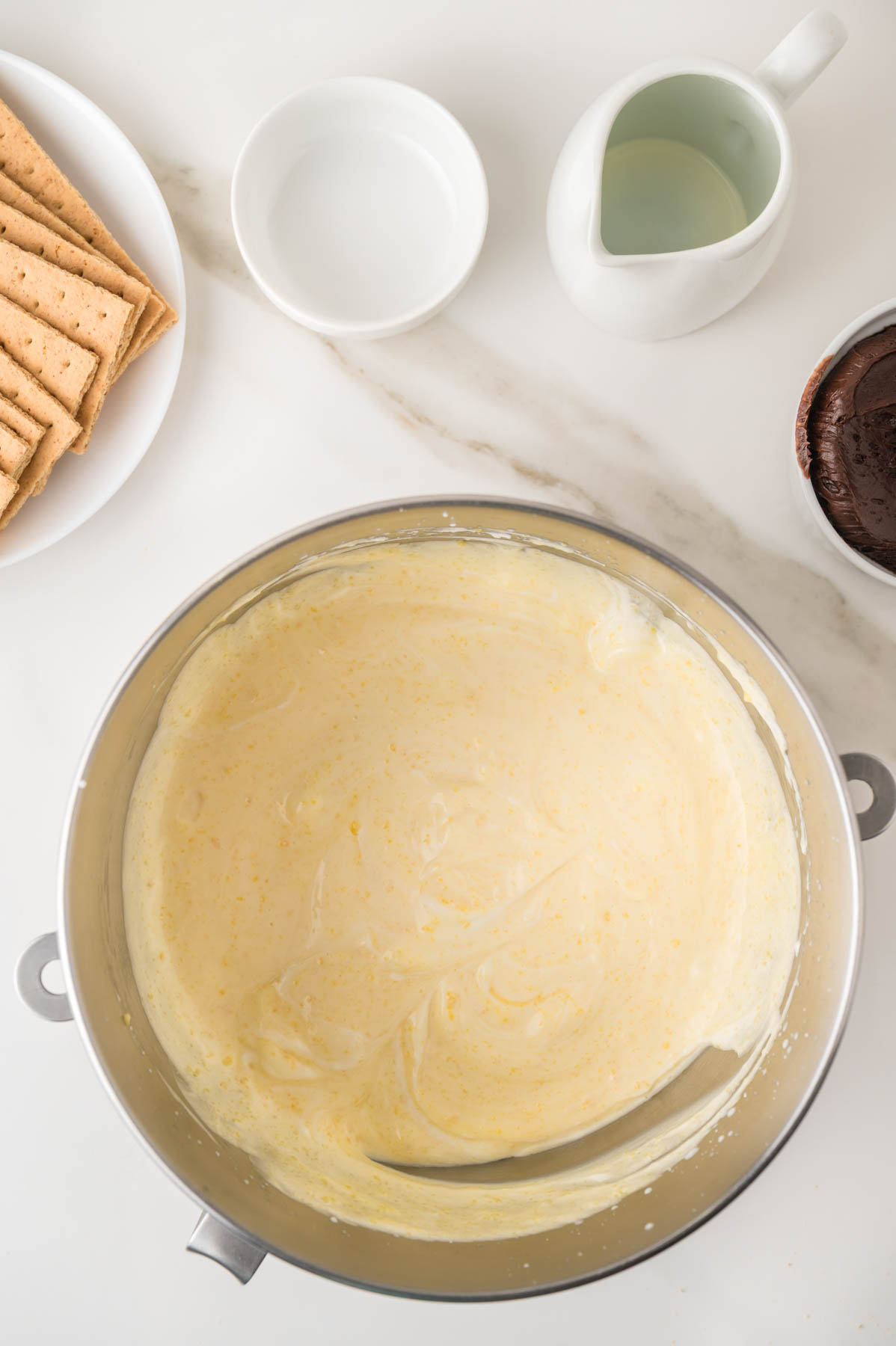 A mixing bowl filled with creamy batter sits on a counter, surrounded by graham crackers, a small pitcher, a white bowl, and a bowl of chocolate.