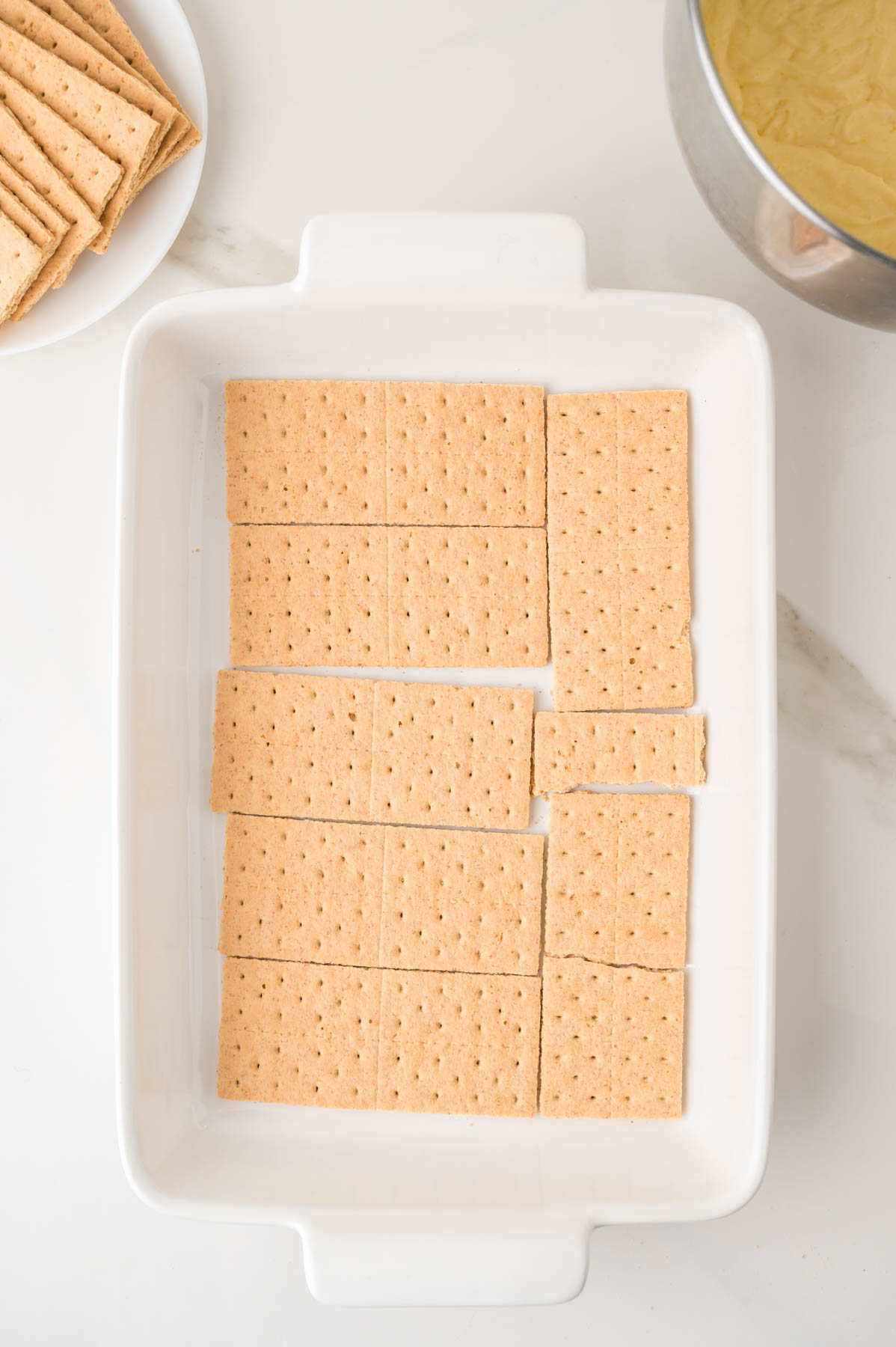 Rectangular pieces of graham crackers are arranged in a single layer at the bottom of a white baking dish, with extra crackers on a plate and a mixing bowl nearby.