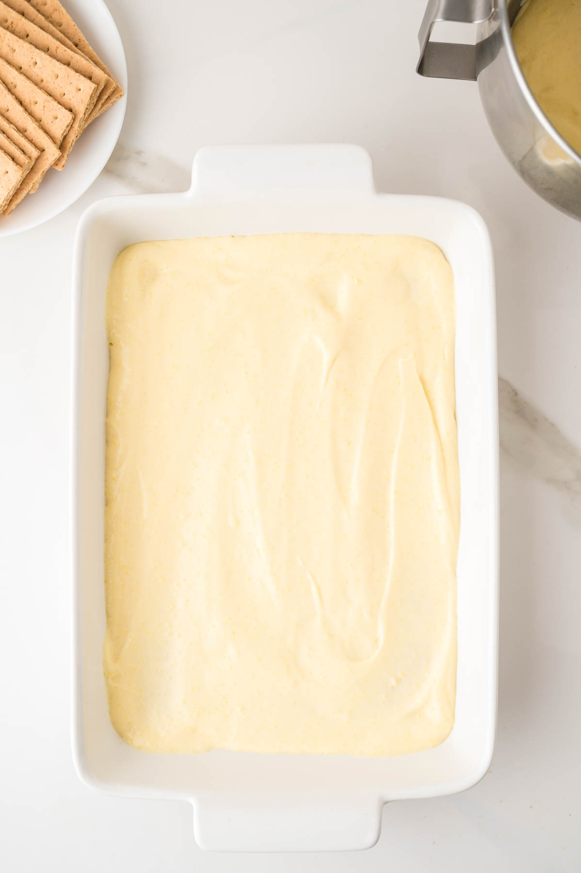 A white baking dish filled with a smooth, pale yellow mixture sits on a marble countertop; graham crackers and a metal mixing bowl are nearby.