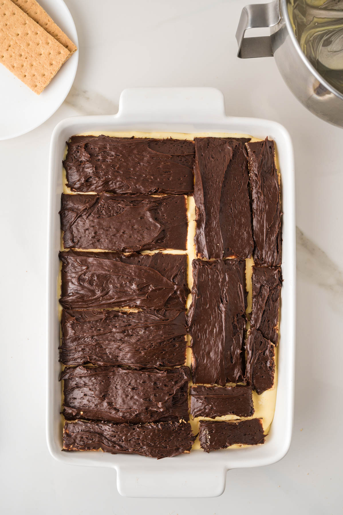 A rectangular white dish filled with layered dessert topped with unevenly spread chocolate frosting, next to a plate with graham crackers and a mixing bowl.
