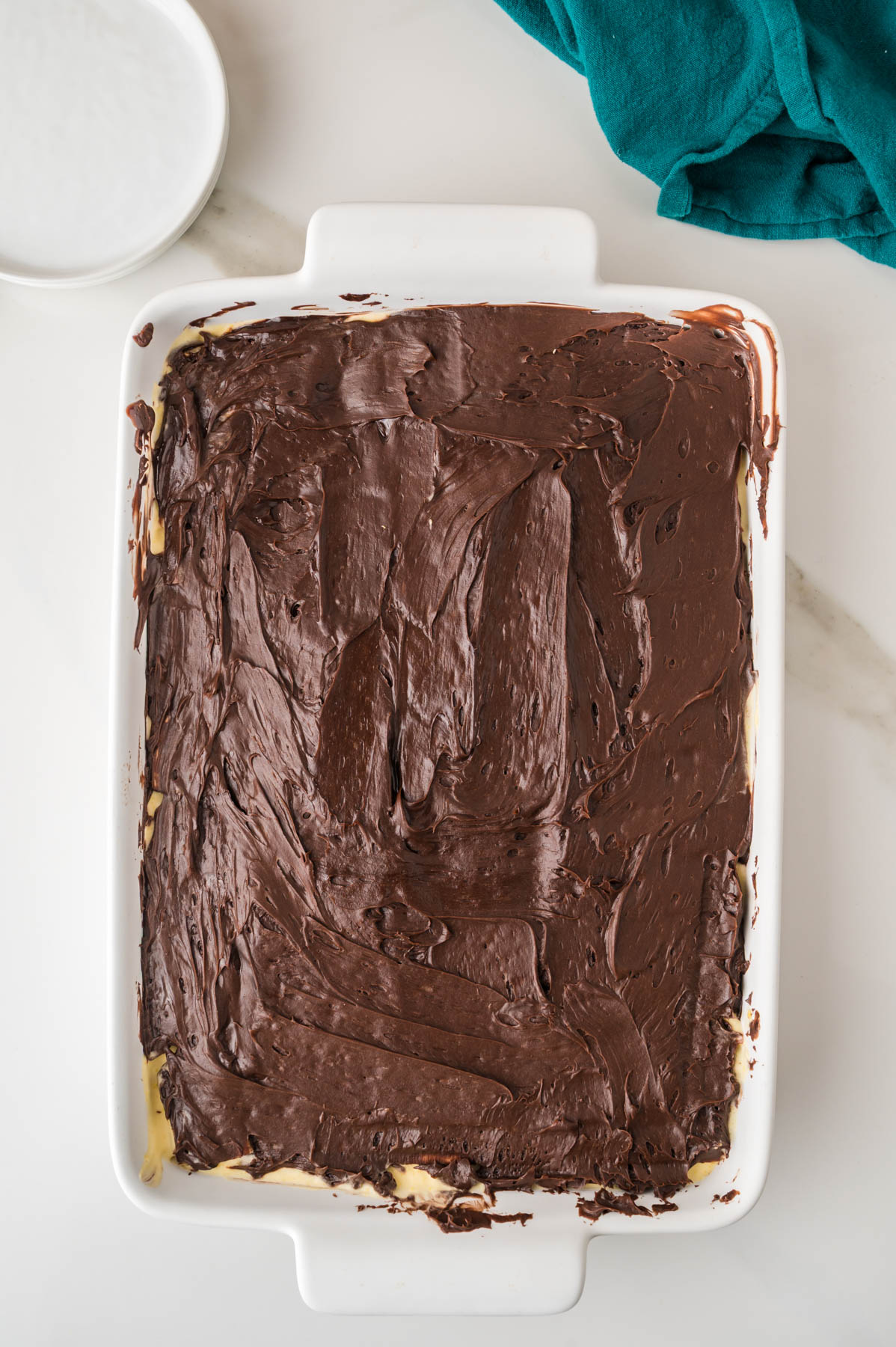 A rectangular white baking dish filled with a dessert topped with a smooth layer of chocolate, on a white surface with a teal cloth and a small white bowl nearby.