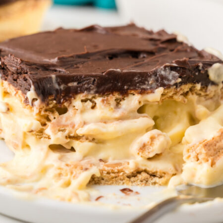A slice of chocolate-topped layered dessert with creamy filling and biscuit layers is served on a white plate with a spoonful taken out.