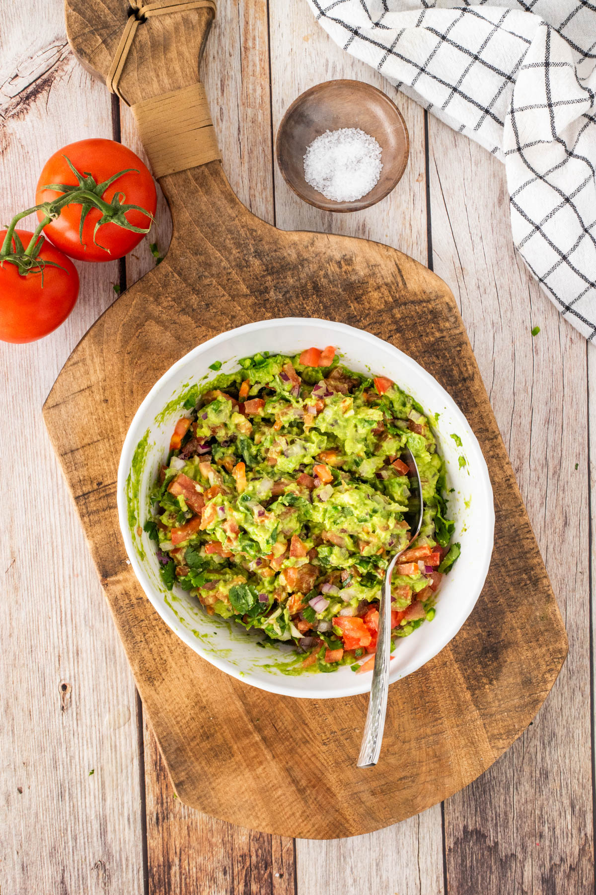 A bowl of guacamole with chopped tomatoes and herbs sits on a wooden board, with a spoon inside, two tomatoes, a small bowl of salt, and a checkered cloth nearby.