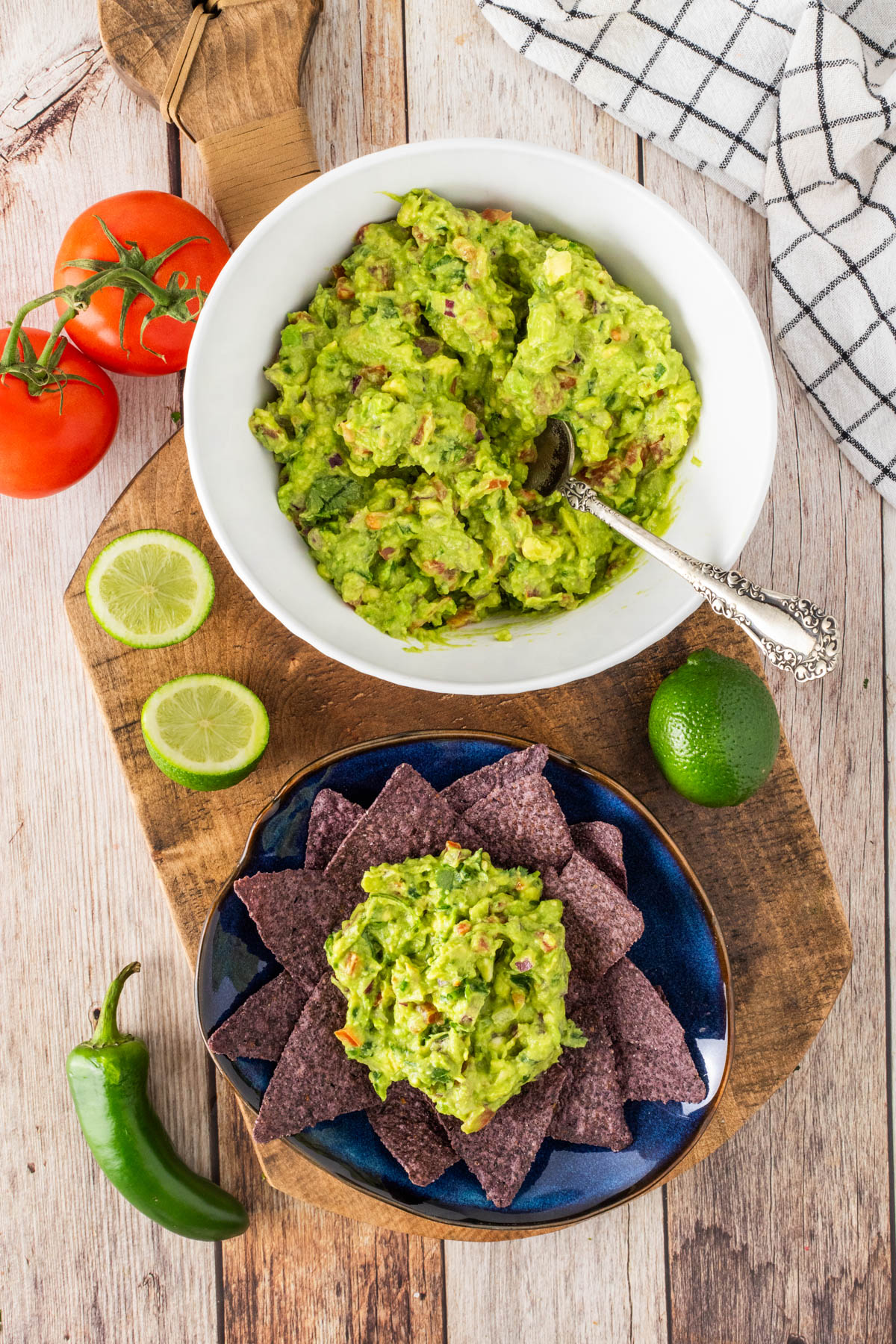 A bowl of guacamole with a spoon, surrounded by fresh tomatoes, lime, a jalape&ntilde;o, and a plate of blue corn tortilla chips topped with guacamole.