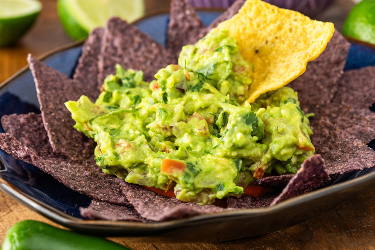 A bowl filled with blue corn tortilla chips topped with a serving of guacamole and a yellow tortilla chip dipped into the guacamole.