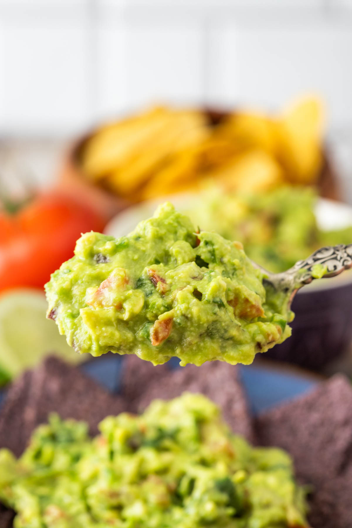 A close-up of a spoonful of guacamole held above a bowl of blue corn chips, with tomatoes, lime, and tortilla chips in the background.
