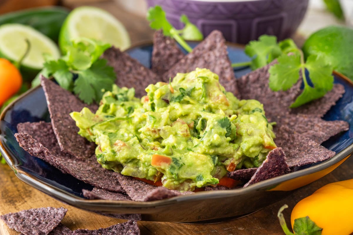 A bowl of guacamole garnished with cilantro, surrounded by blue corn tortilla chips, with lime slices and herbs in the background.