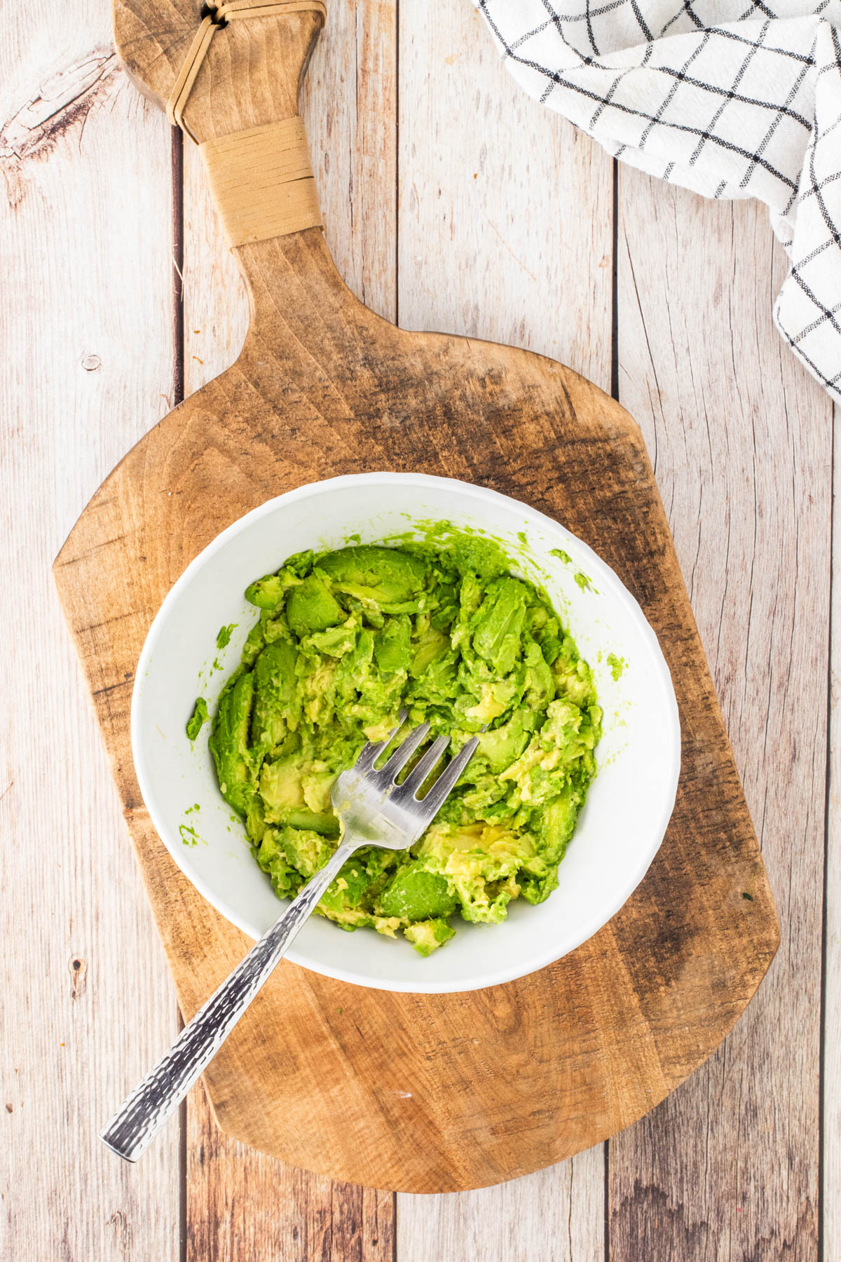 A white bowl of mashed avocado with a fork rests on a wooden cutting board, with part of a checkered cloth visible in the corner.