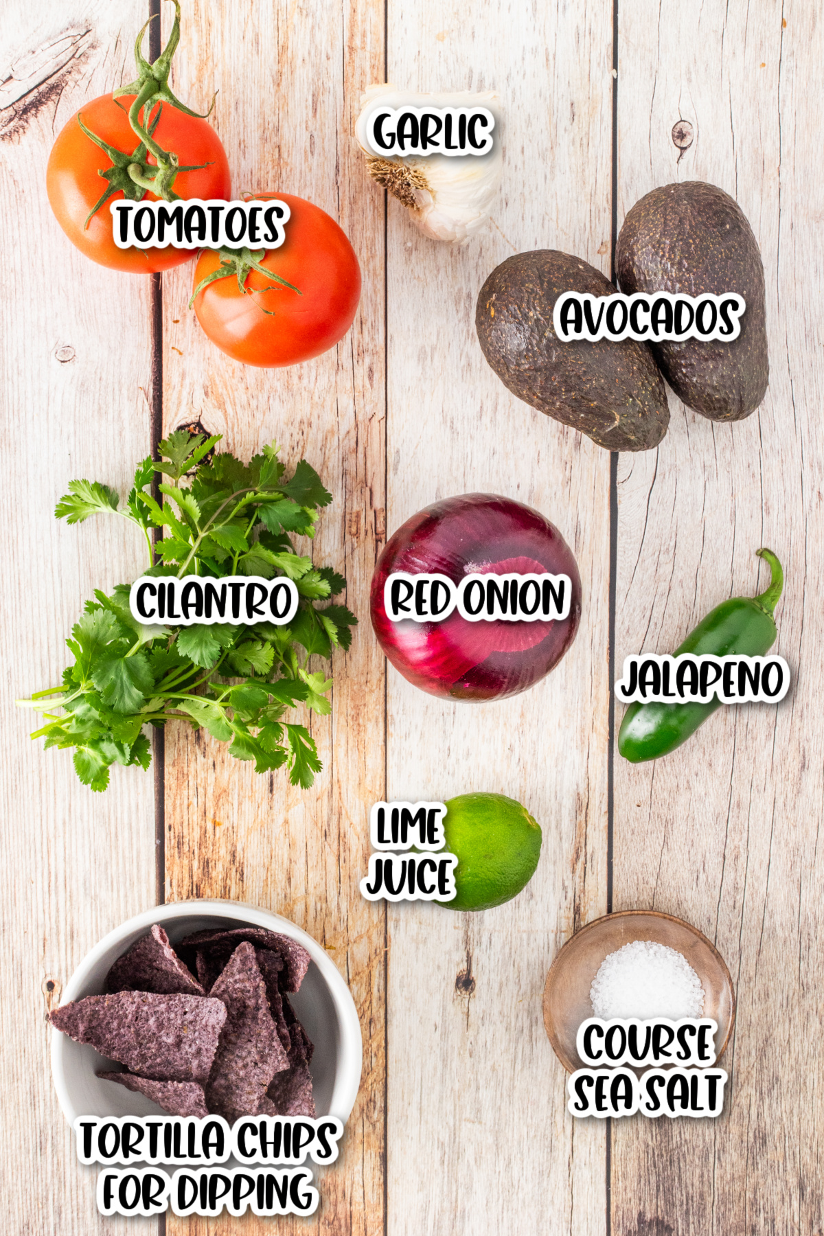 Ingredients for guacamole including tomatoes, garlic, avocados, cilantro, red onion, jalapeno, lime juice, course sea salt, and tortilla chips arranged on a wooden surface.