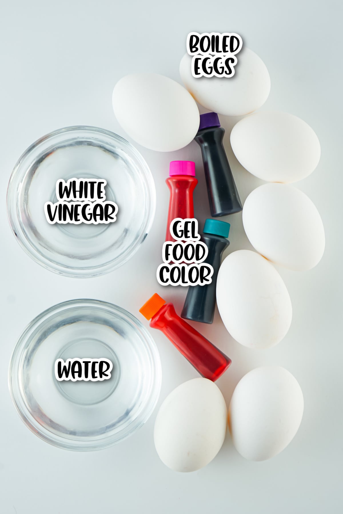 A flat lay of boiled eggs, bowls of white vinegar and water, and three bottles of gel food color on a white surface.