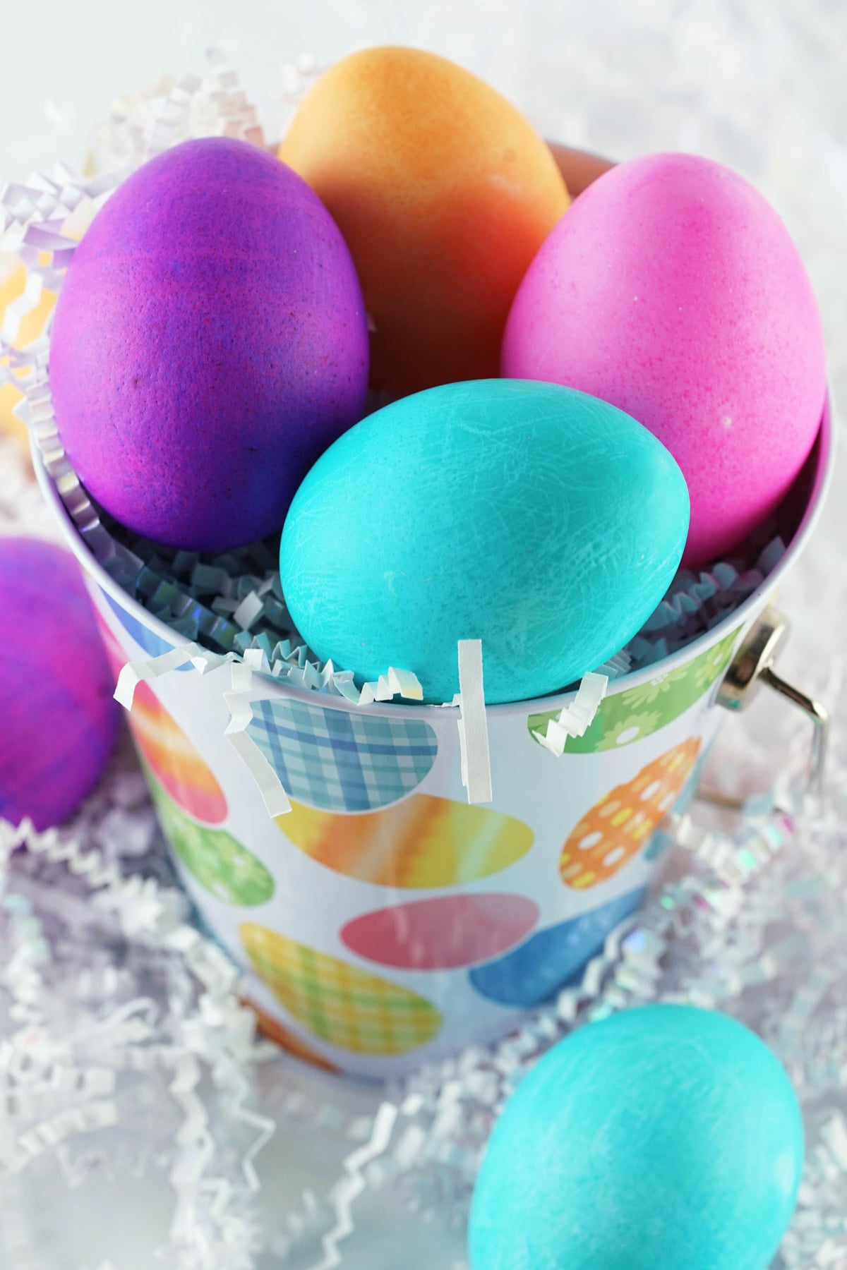 A small bucket decorated with colorful patterns holds brightly dyed Easter eggs in purple, blue, orange, and pink, surrounded by white paper shreds.