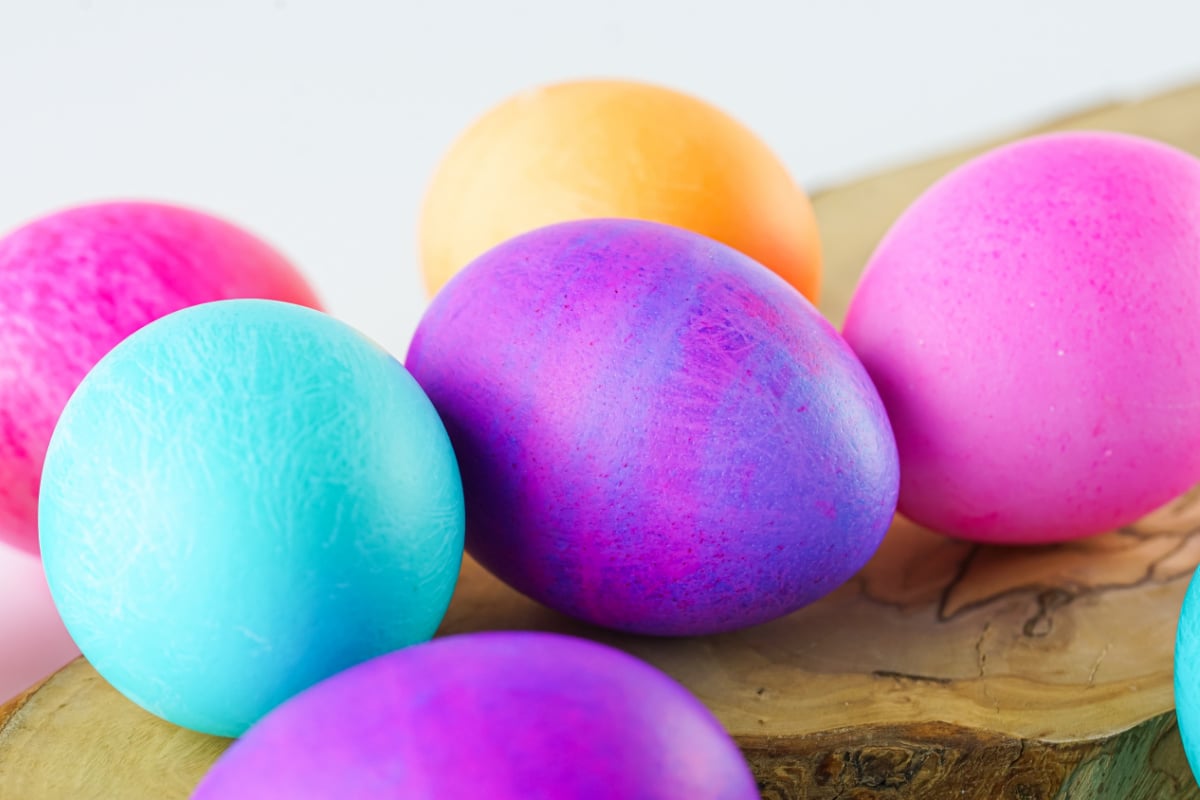 Several brightly colored Easter eggs, including purple, pink, blue, and orange, are arranged on a wooden surface against a light background.