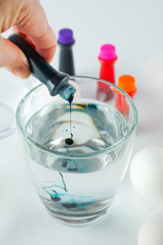 A hand holds a dropper releasing blue food coloring into a glass of clear water, with bottles of food coloring and white eggs in the background.