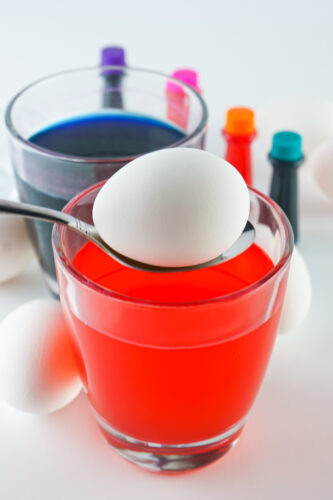 A white egg on a spoon is held above a glass filled with red liquid dye, with another glass of blue dye and bottles of food coloring in the background.