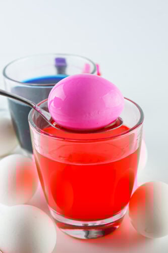 A pink-dyed egg rests on a spoon above a glass of red dye, with a glass of blue dye and several white eggs nearby.