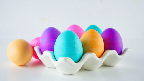 A white ceramic egg tray holds six colorful dyed eggs, with one light orange egg resting outside the tray on a white surface.