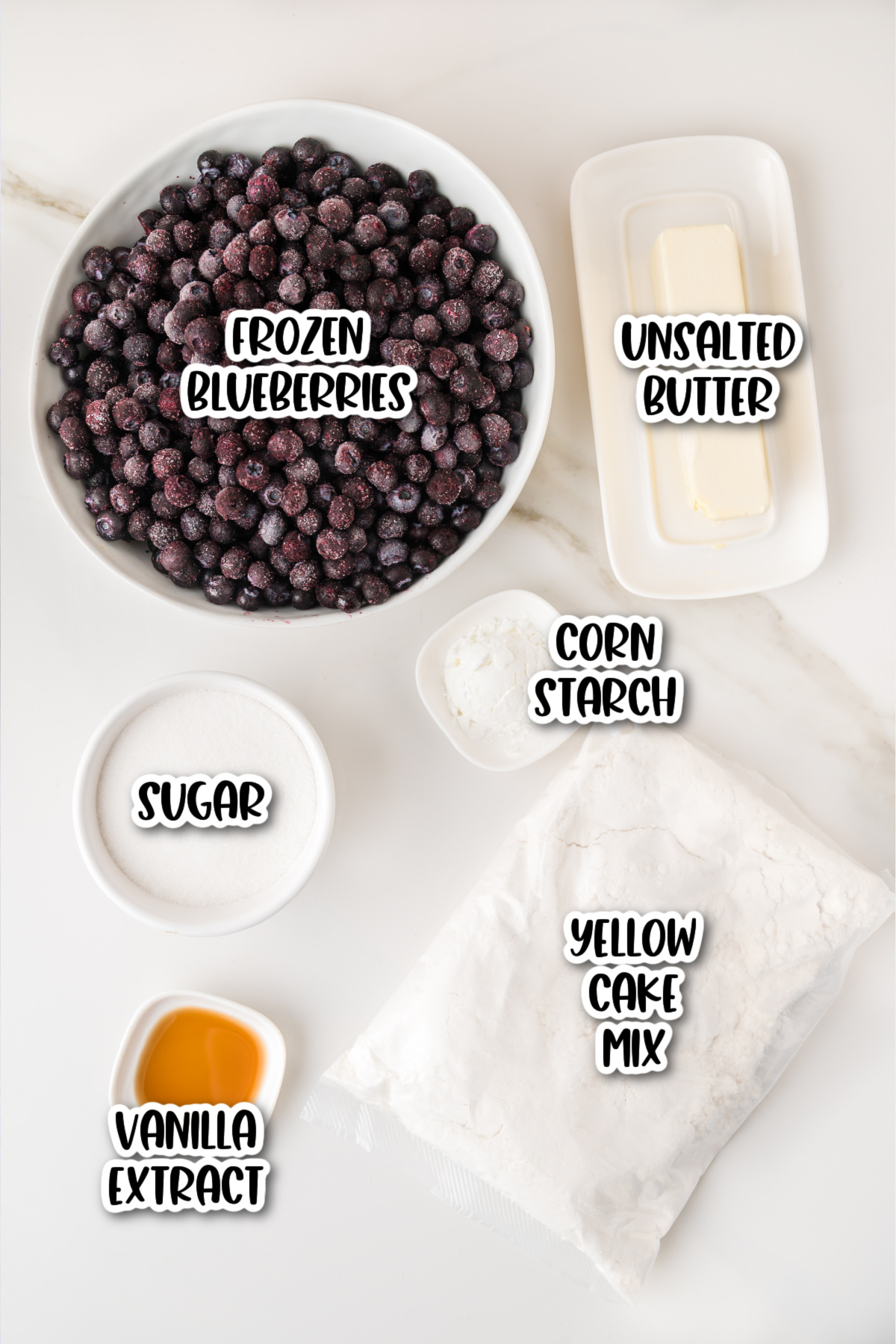 A bowl of frozen blueberries, a stick of unsalted butter, sugar, cornstarch, yellow cake mix, and vanilla extract arranged on a white surface.