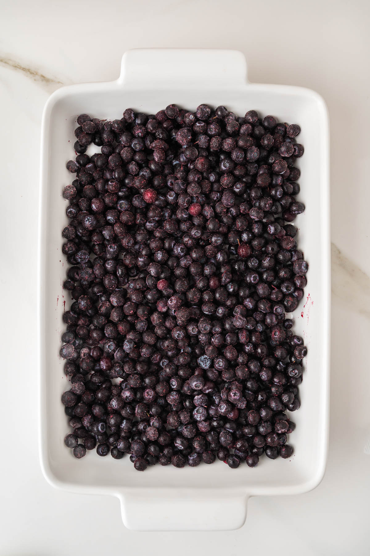 A white rectangular dish filled with a single layer of frozen blueberries on a light-colored surface.