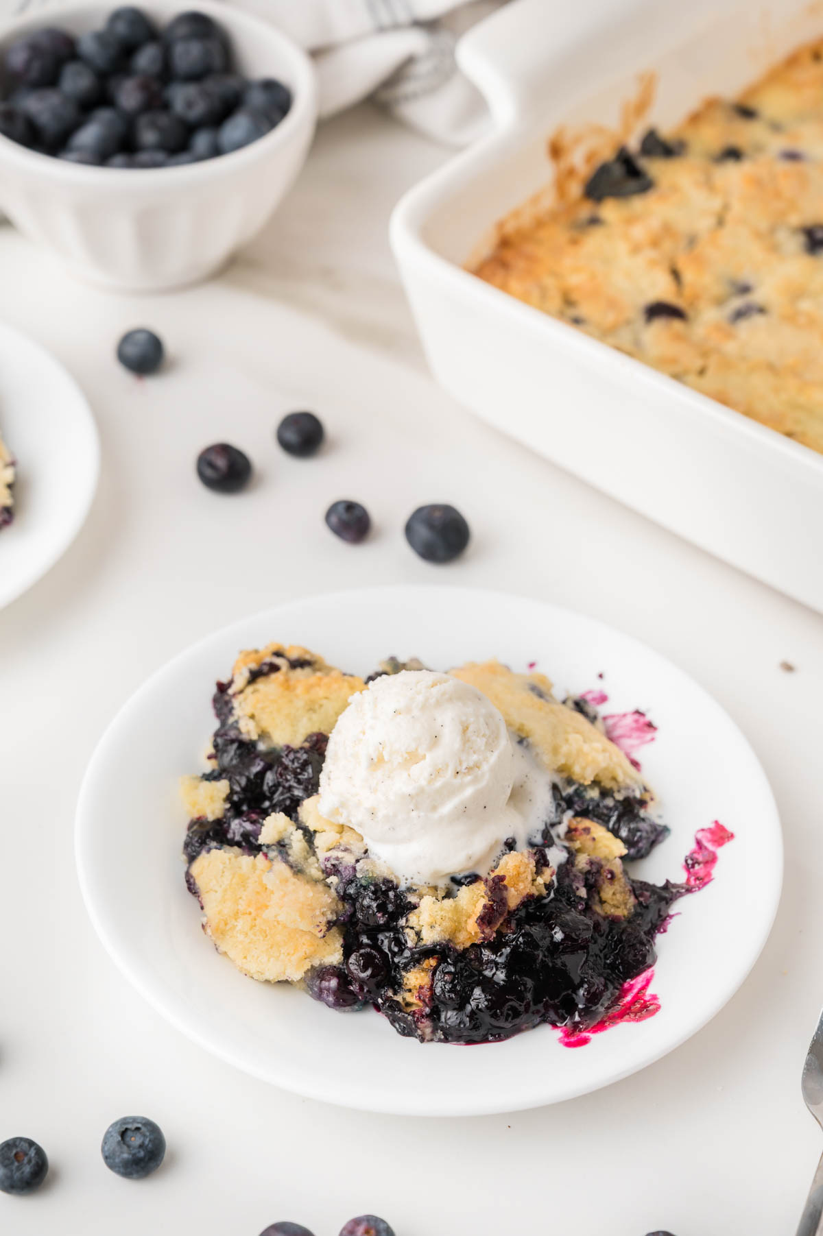 A serving of blueberry cobbler topped with a scoop of vanilla ice cream sits on a white plate, with fresh blueberries and a baking dish in the background.