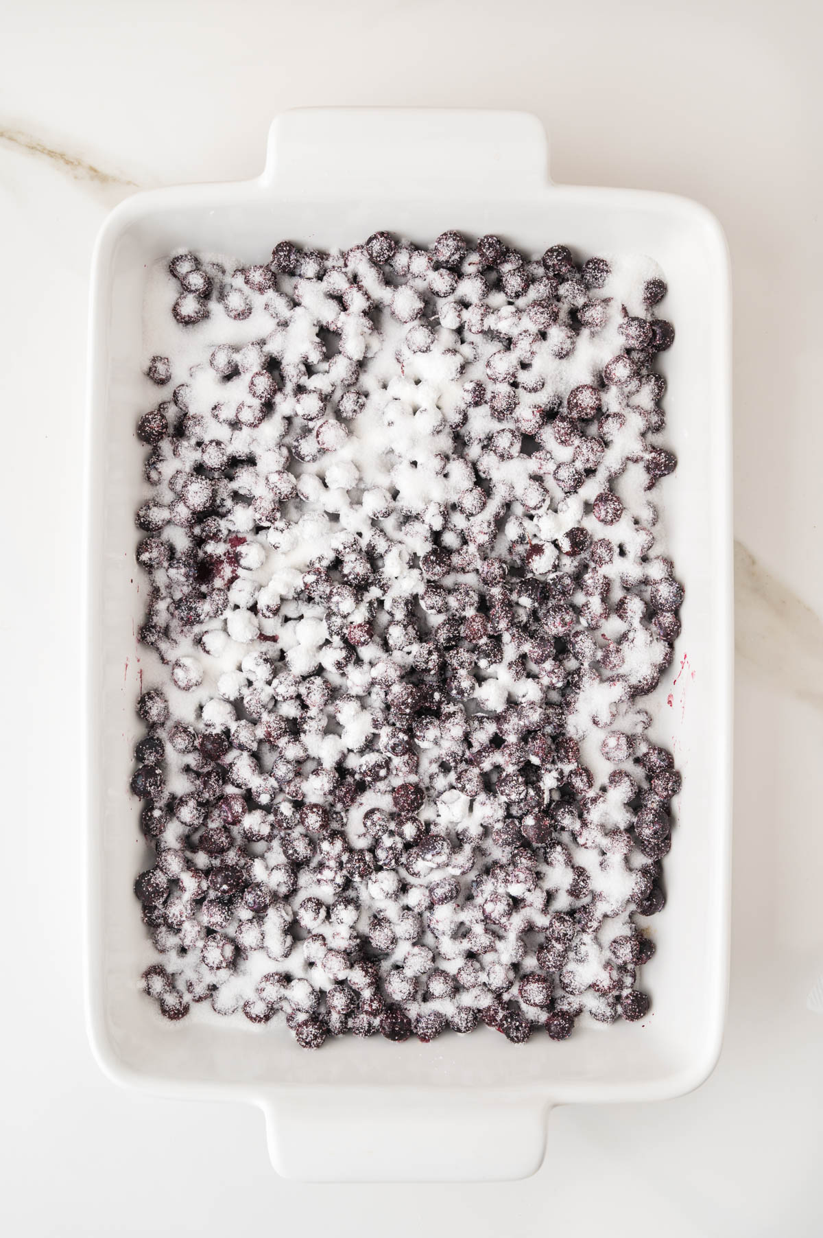 A white baking dish filled with blueberries covered in a layer of granulated sugar, placed on a light-colored surface.