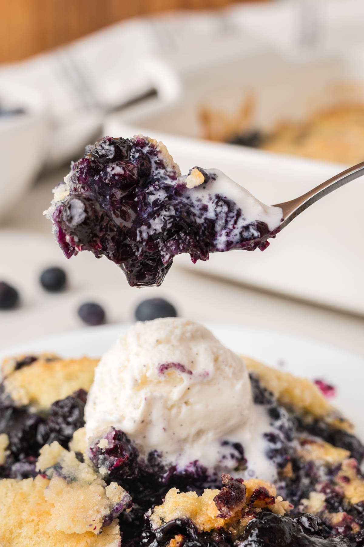 A close-up of a blueberry cobbler topped with a scoop of vanilla ice cream; a fork holds a bite of cobbler above the plate.