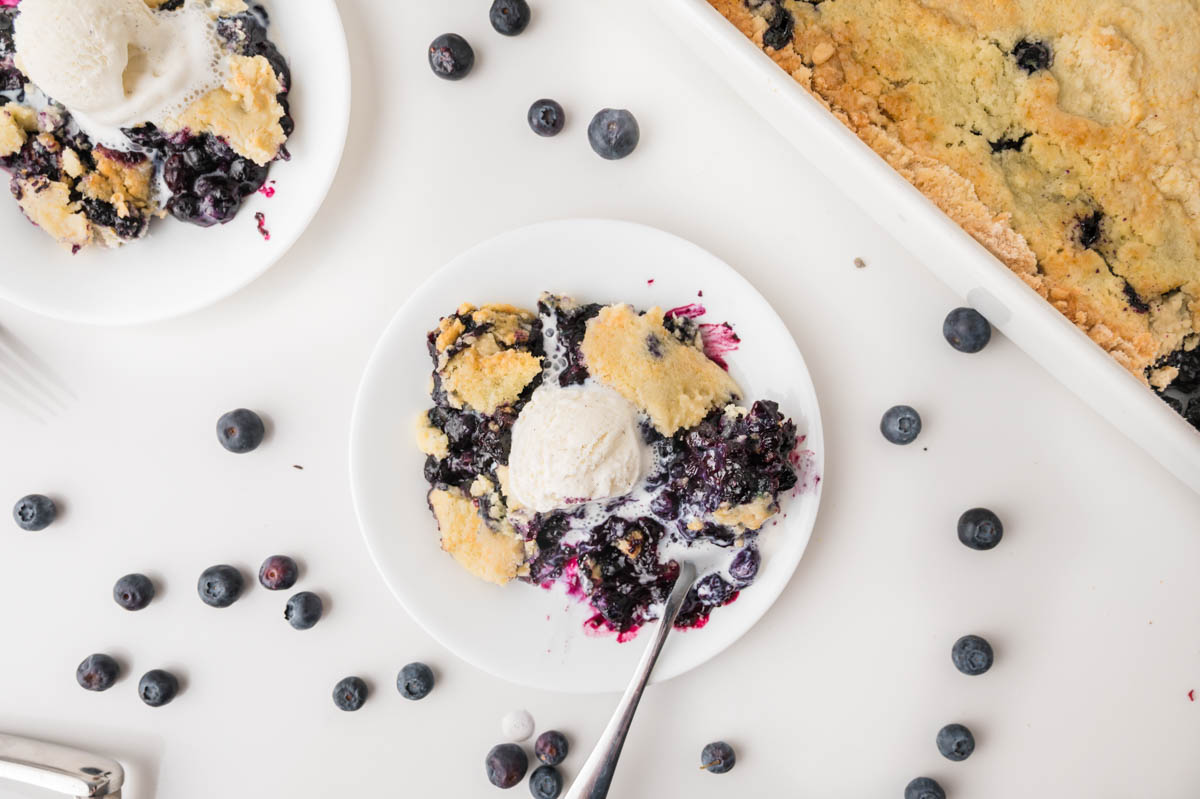 A plate of blueberry cobbler topped with a scoop of vanilla ice cream, surrounded by fresh blueberries and a baking dish with more cobbler on a white surface.