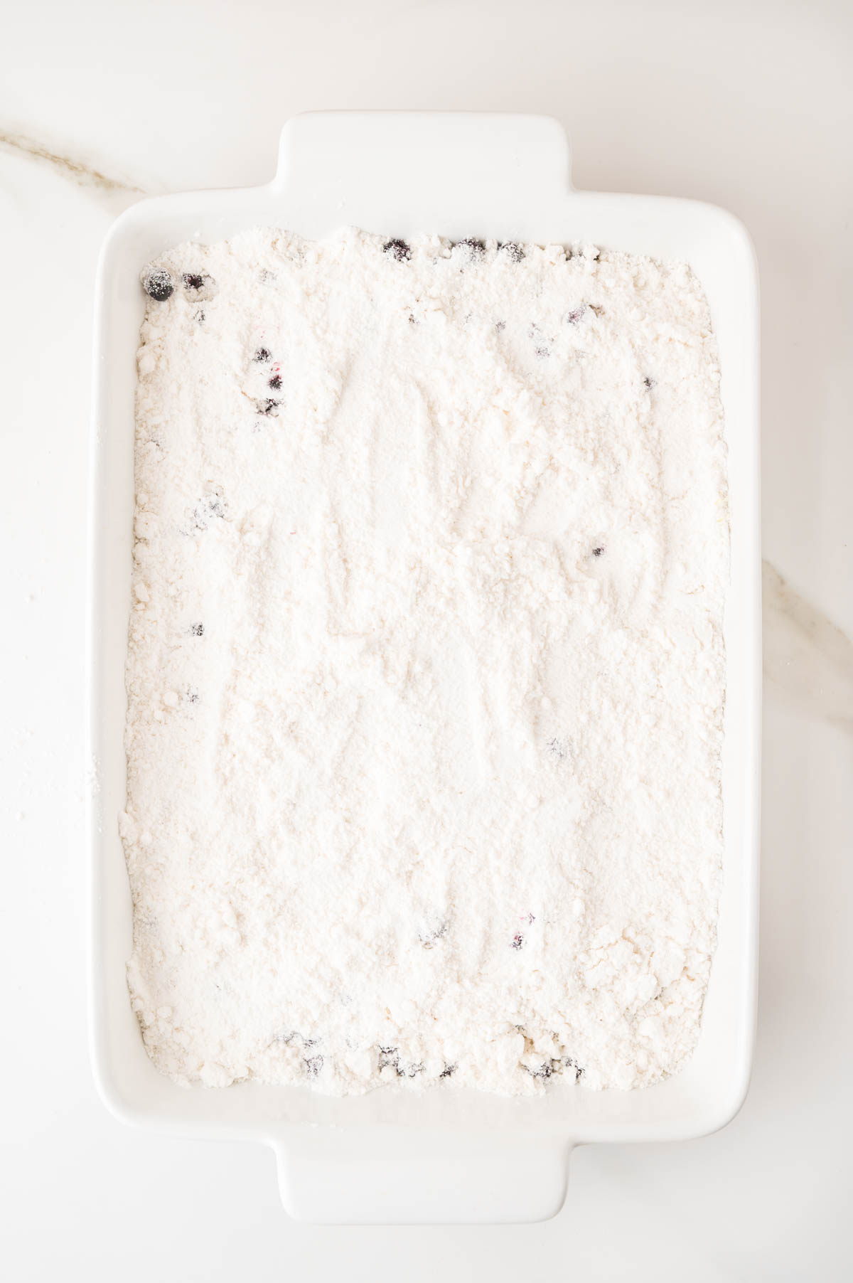 A white rectangular baking dish filled with a layer of yellow cake mix with visible blueberries, set on a white surface.