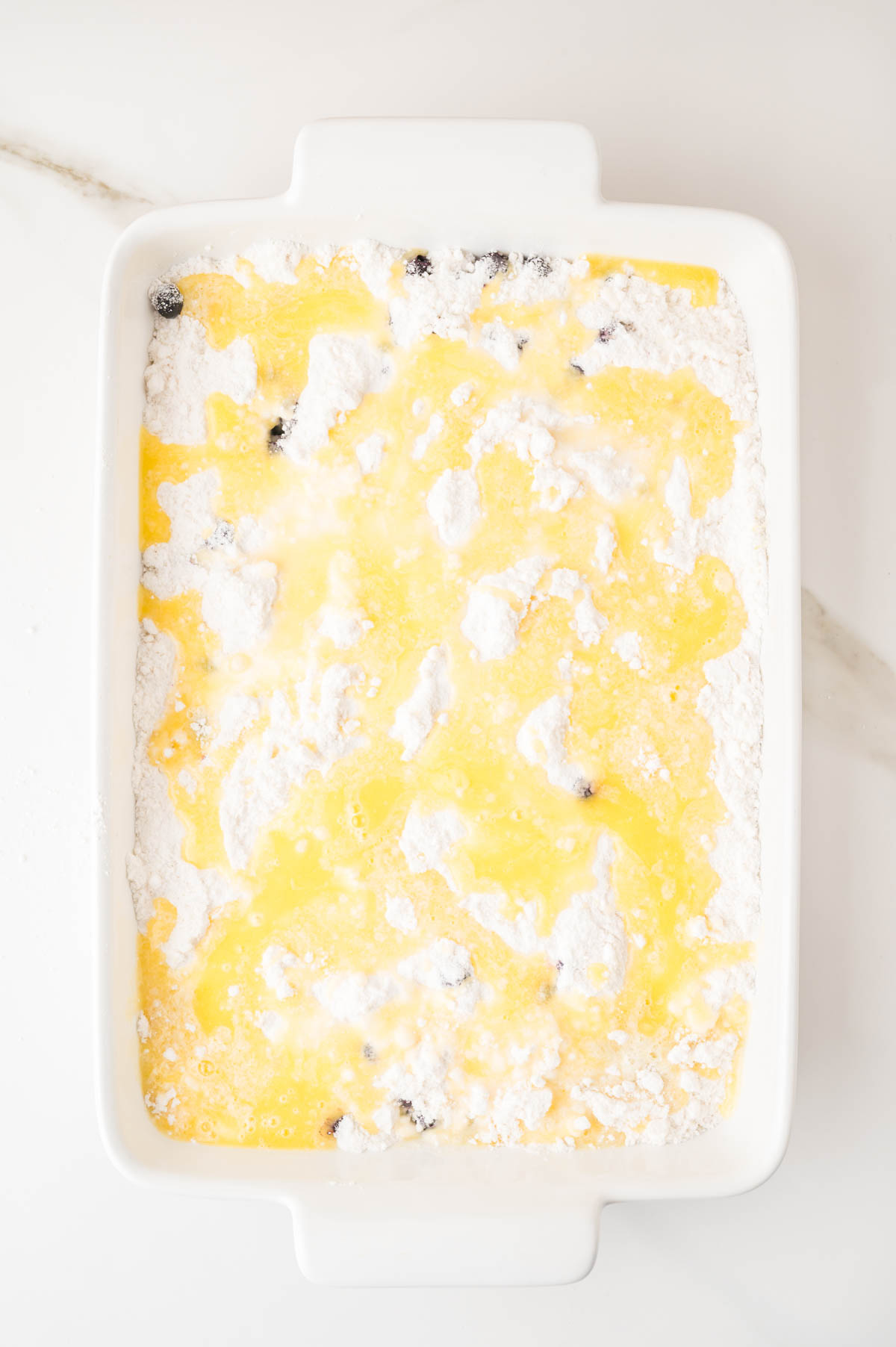Rectangular white baking dish filled with cake mix and melted butter mixture, with visible blueberries, on a white countertop.