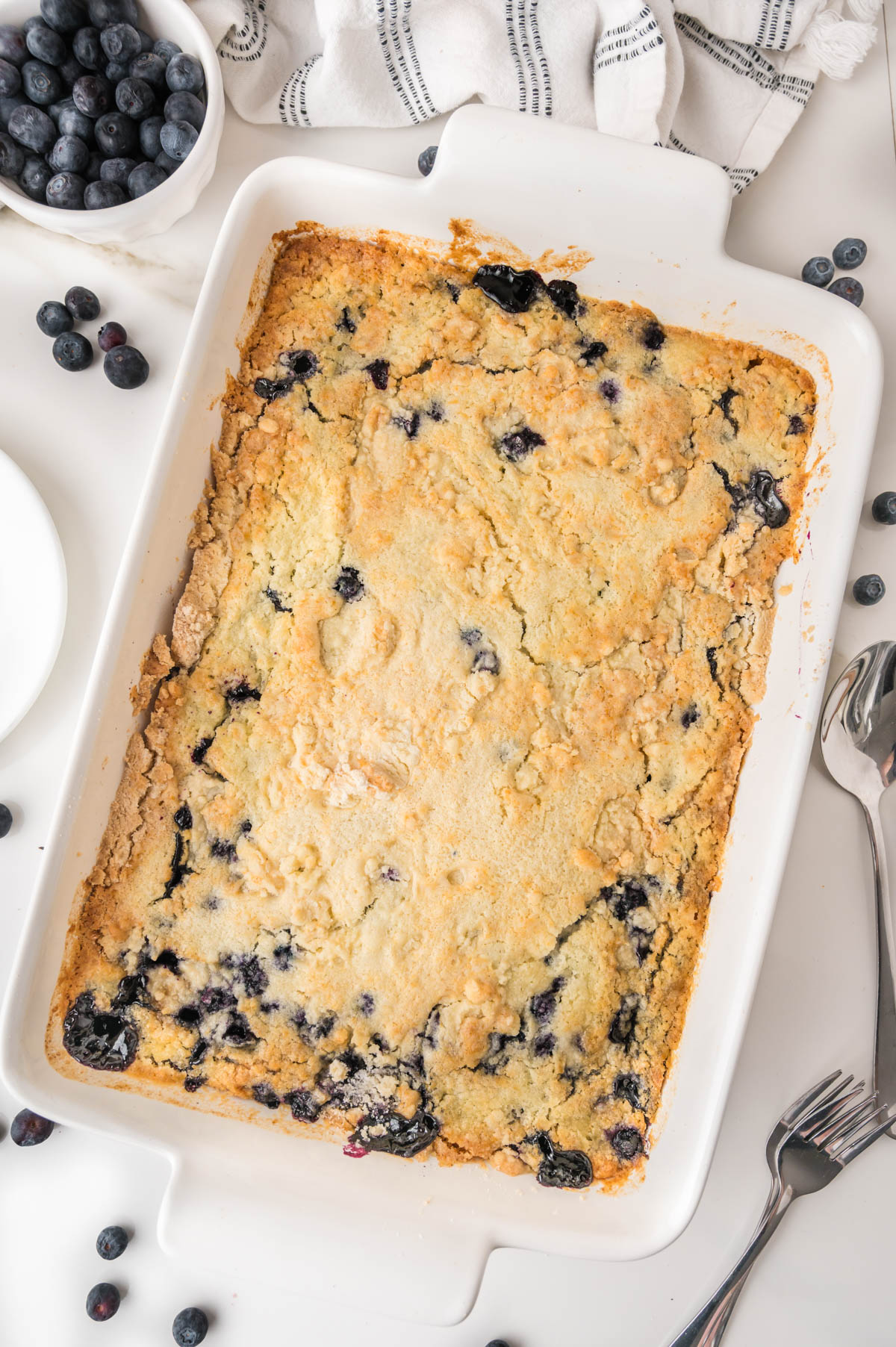 A baked blueberry cobbler in a white rectangular dish, surrounded by fresh blueberries, plates, utensils, and a striped towel on a white surface.