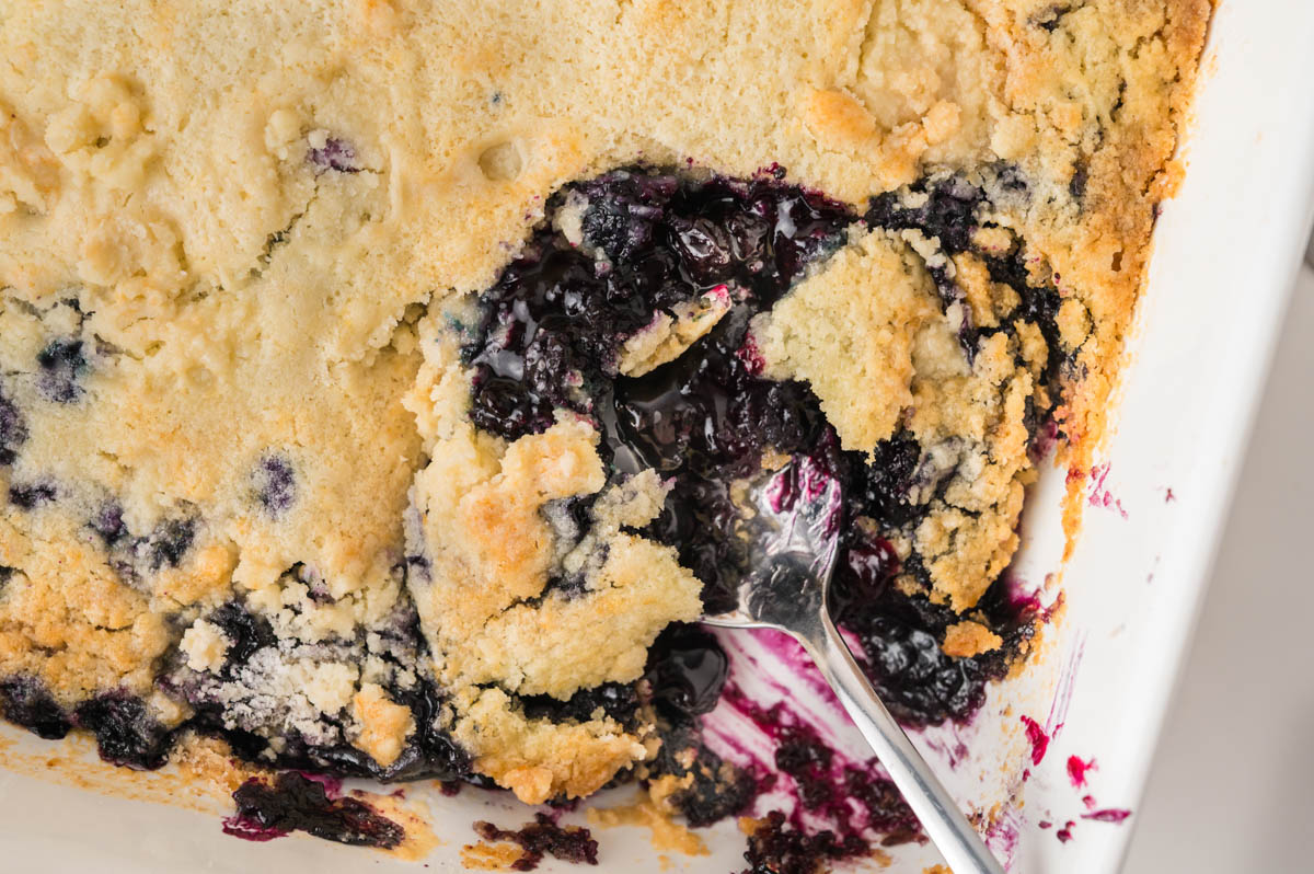A close-up of a partially served blueberry dump cake in a white baking dish, with a metal spoon scooping out a portion.