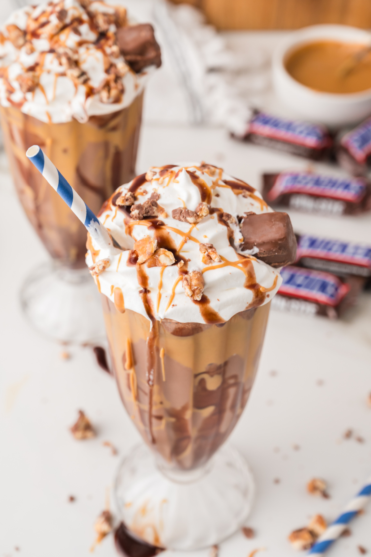 Two chocolate milkshakes topped with whipped cream, caramel drizzle, and chopped candy bars, served in tall glasses with striped straws. Snickers bars are visible in the background.