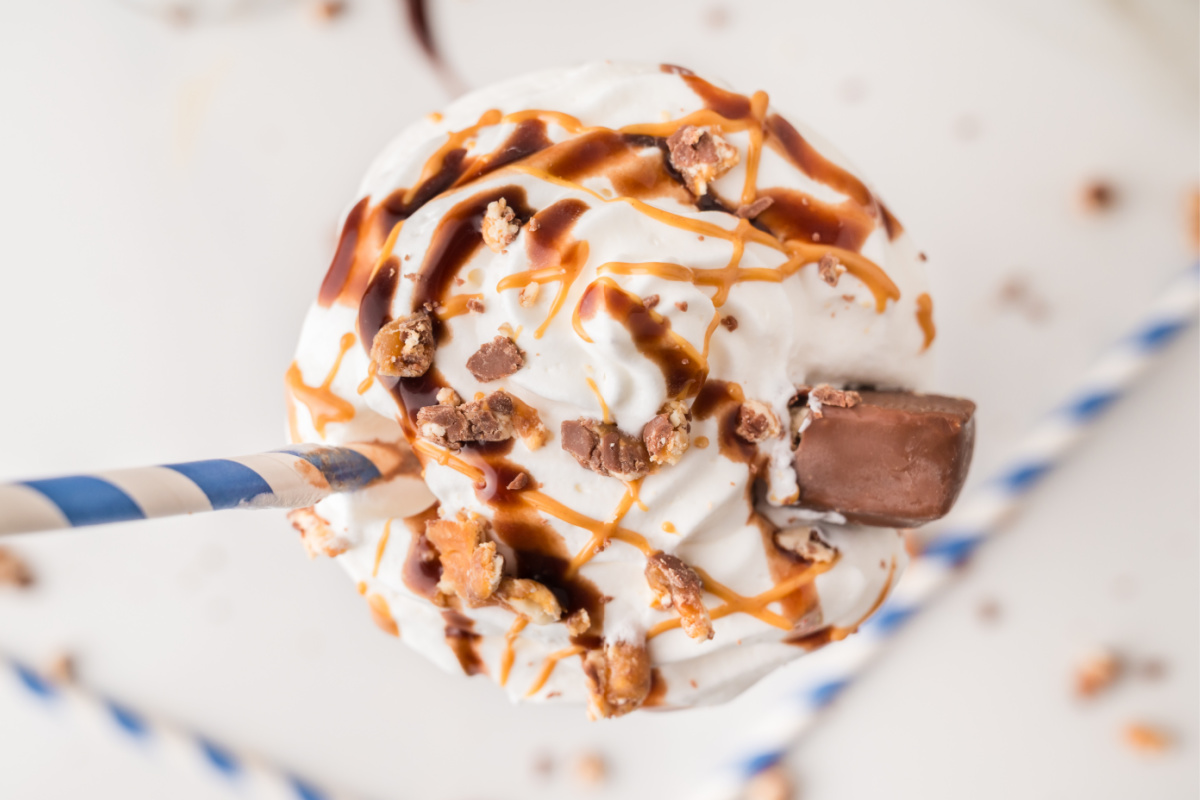 A close-up of a whipped cream-topped milkshake with caramel and chocolate drizzle, crushed candy pieces, a chocolate bar, and a blue-striped straw.
