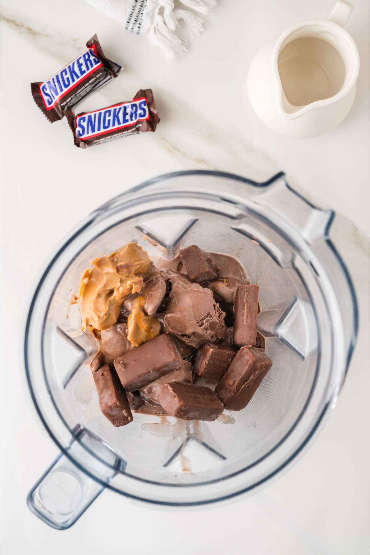 A blender contains Snickers bars, chocolate ice cream, and peanut butter. Two wrapped Snickers bars and a small pitcher of milk are on the counter nearby.