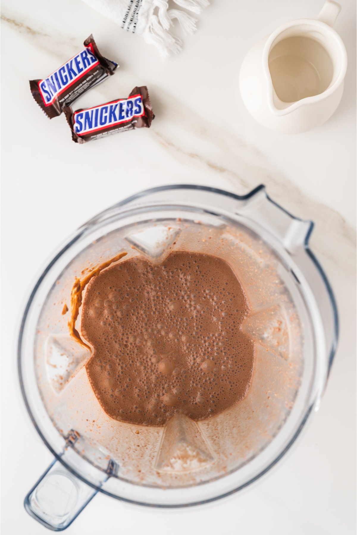 A blender with chocolate mixture inside sits on a counter next to two Snickers bars and a small white pitcher.