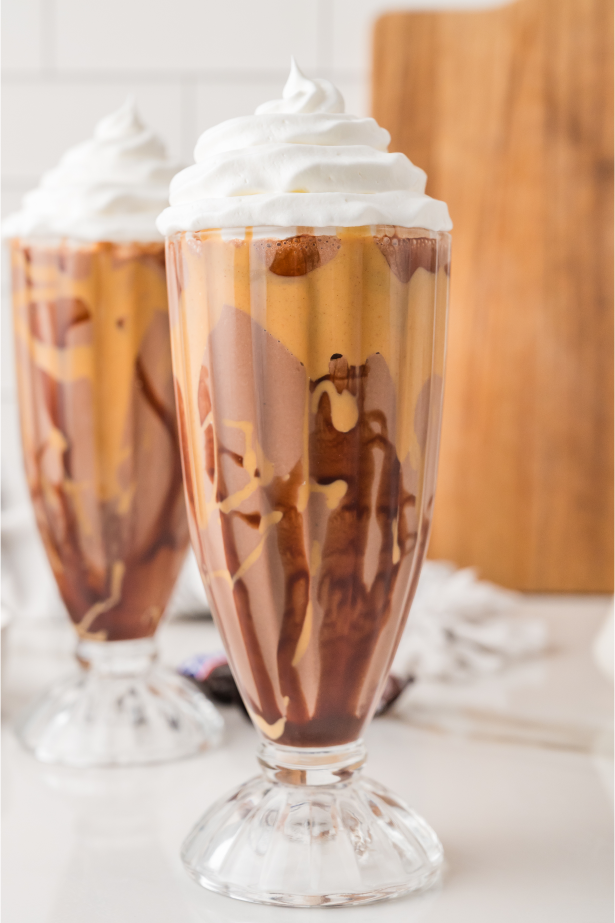 Two Snickers milkshakes in tall glass cups, topped with whipped cream, on a white counter with a wooden board in the background.