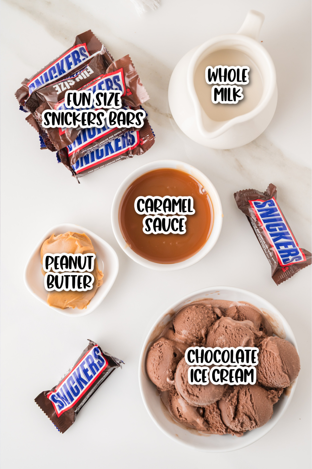 Top-down view of a small pitcher of milk, caramel sauce, peanut butter, chocolate ice cream, and fun size Snickers bars arranged on a white surface.