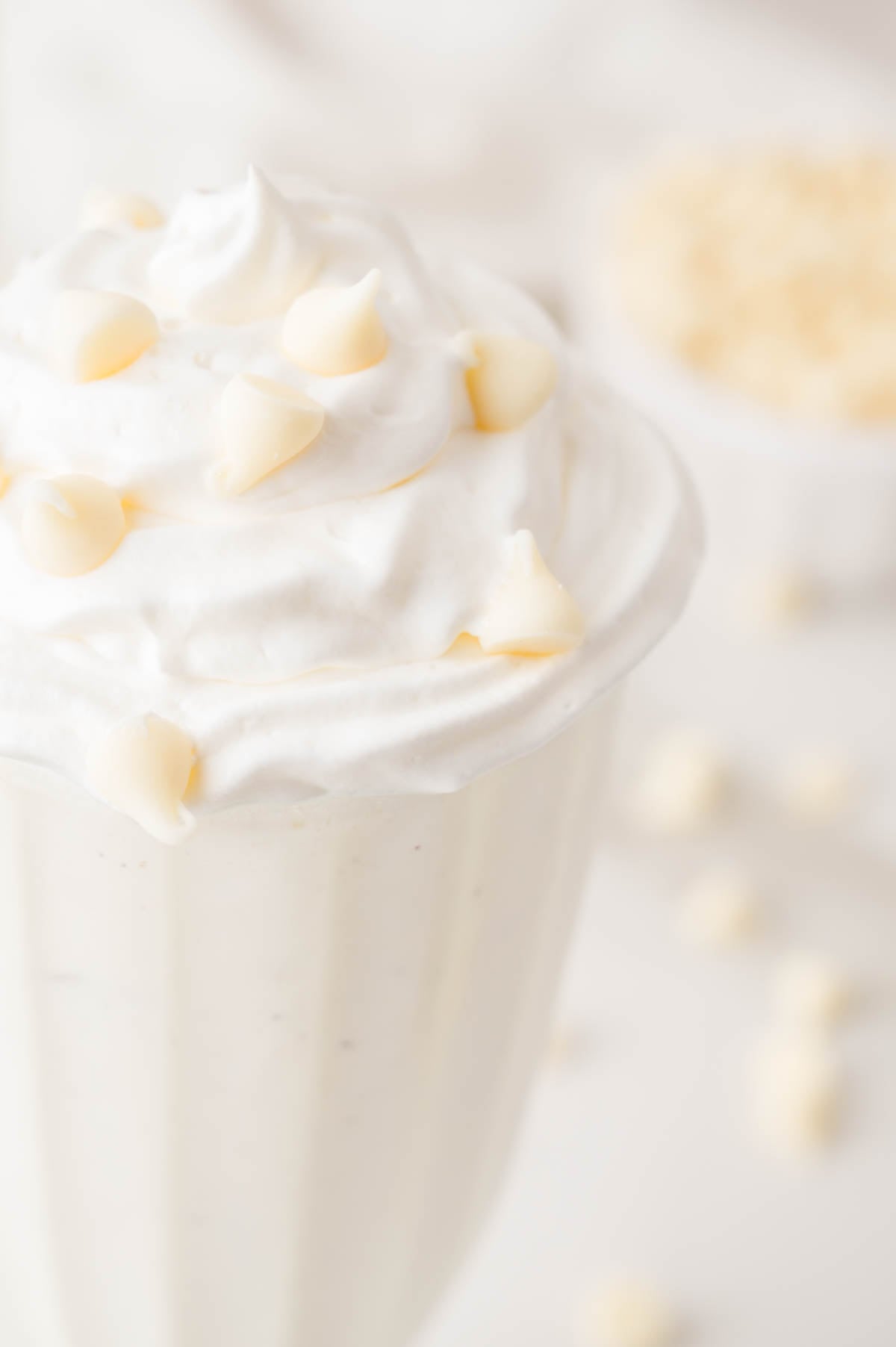 A glass of creamy white milkshake topped with whipped cream and white chocolate chips, with more chocolate chips scattered in the background.