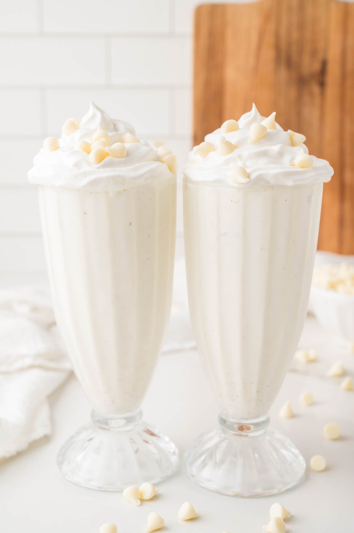 Two tall glasses of vanilla milkshake topped with whipped cream and white chocolate chips, placed on a white surface with scattered white chocolate chips.