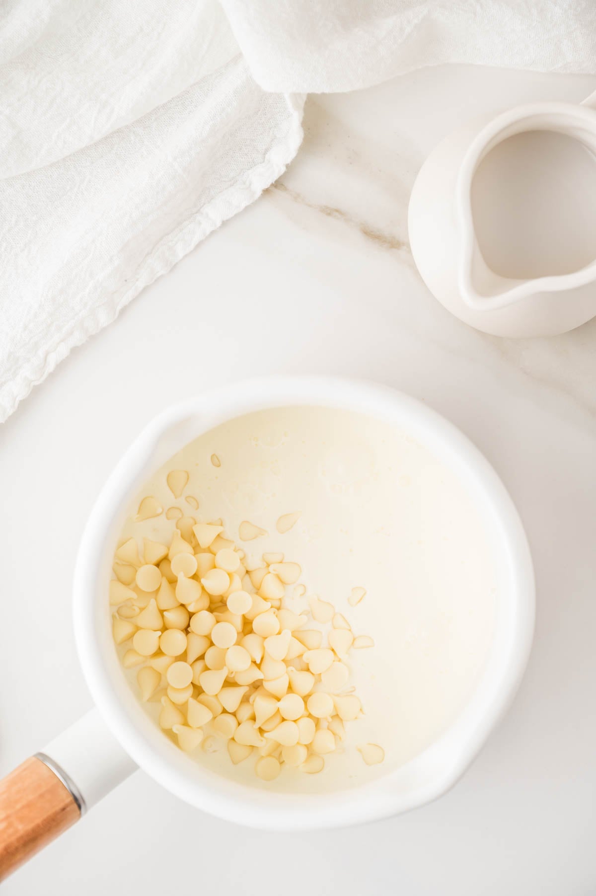 A white saucepan filled with cream and white chocolate chips sits on a marble surface beside a small white pitcher and a folded white cloth.
