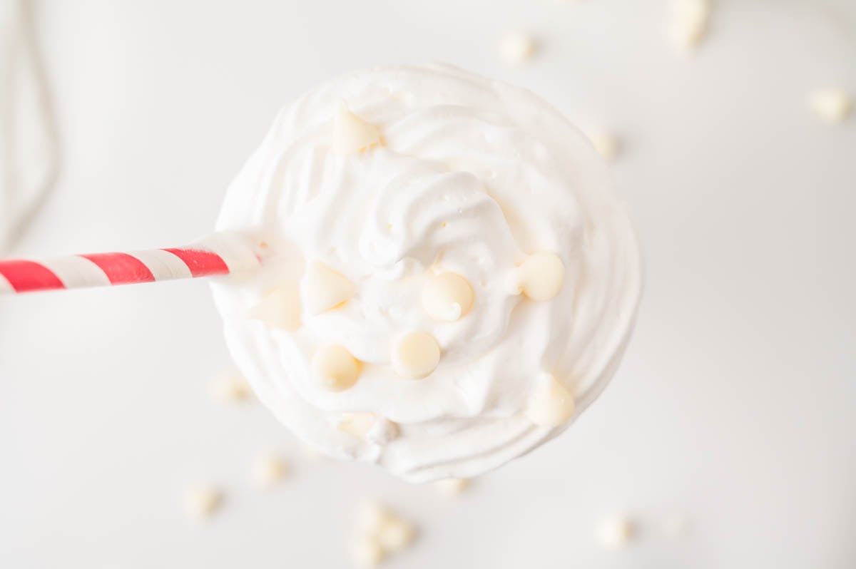 A top-down view of a whipped creamy white beverage with white chocolate chips and a red-striped straw on a white background.