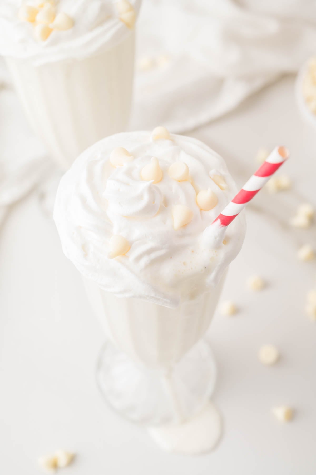 A glass of white milkshake topped with whipped cream and white chocolate chips, served with a red and white striped straw.