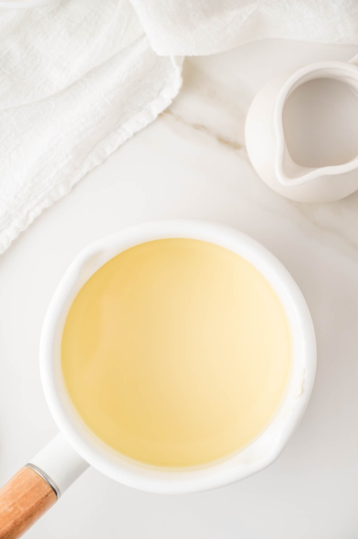 A white saucepan filled with a pale yellow liquid sits on a white surface next to a small white pitcher and a folded white cloth.