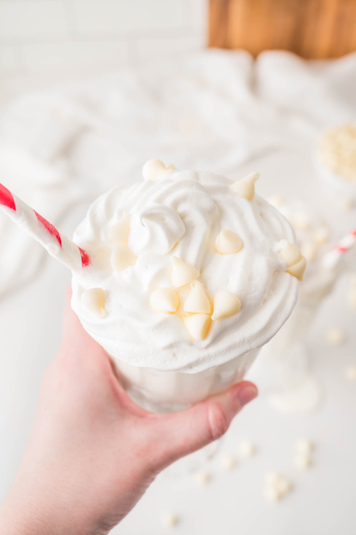 A hand holds a glass of milkshake topped with whipped cream and white chocolate chips, with a red-striped straw.